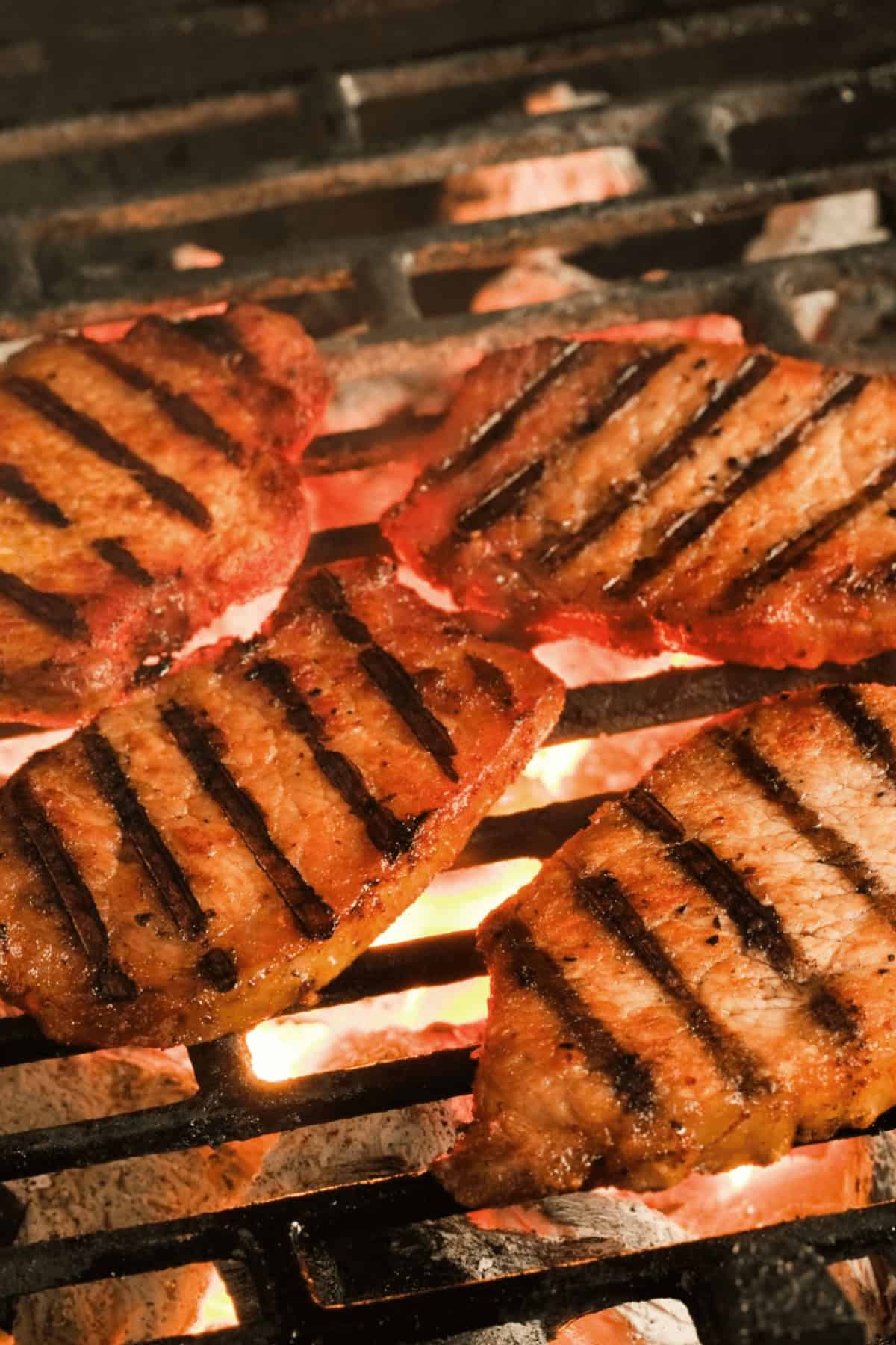 Four grilled pork chops with char marks are cooking on a barbecue grill over hot coals, their savory marinade wafting through the air.