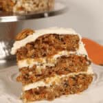 A slice of carrot cake with pineapple, coconut, and cream cheese frosting and a pecan on top, displayed on a white plate.