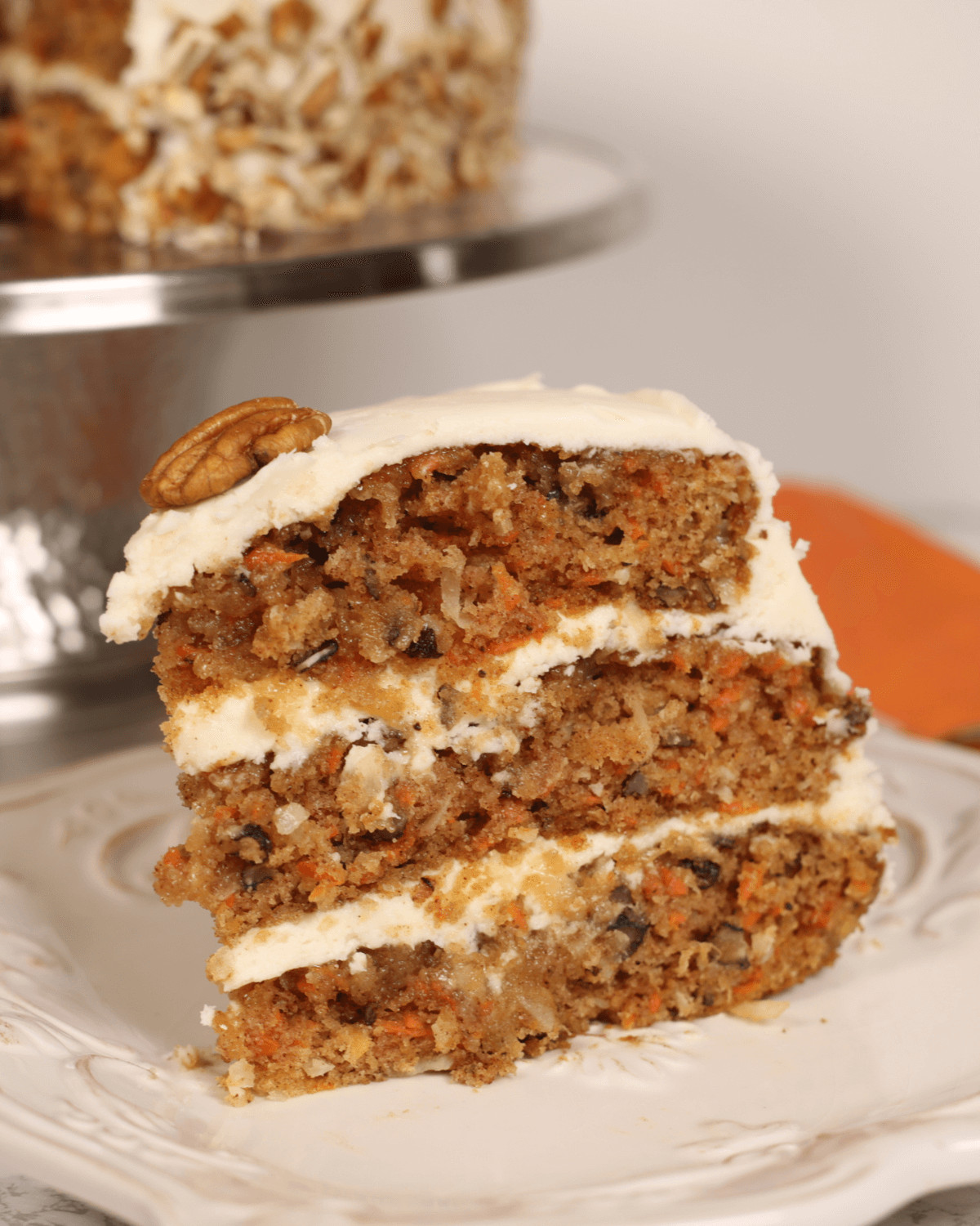 A slice of carrot cake with pineapple, coconut, and cream cheese frosting and a pecan on top, displayed on a white plate.