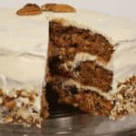 A carrot cake with pineapple and coconut, cream cheese frosting, and chopped nuts, with one slice removed.