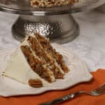 A slice of carrot cake with pineapple, coconut, and cream cheese frosting on a plate, with a cake stand and more cake in the background.