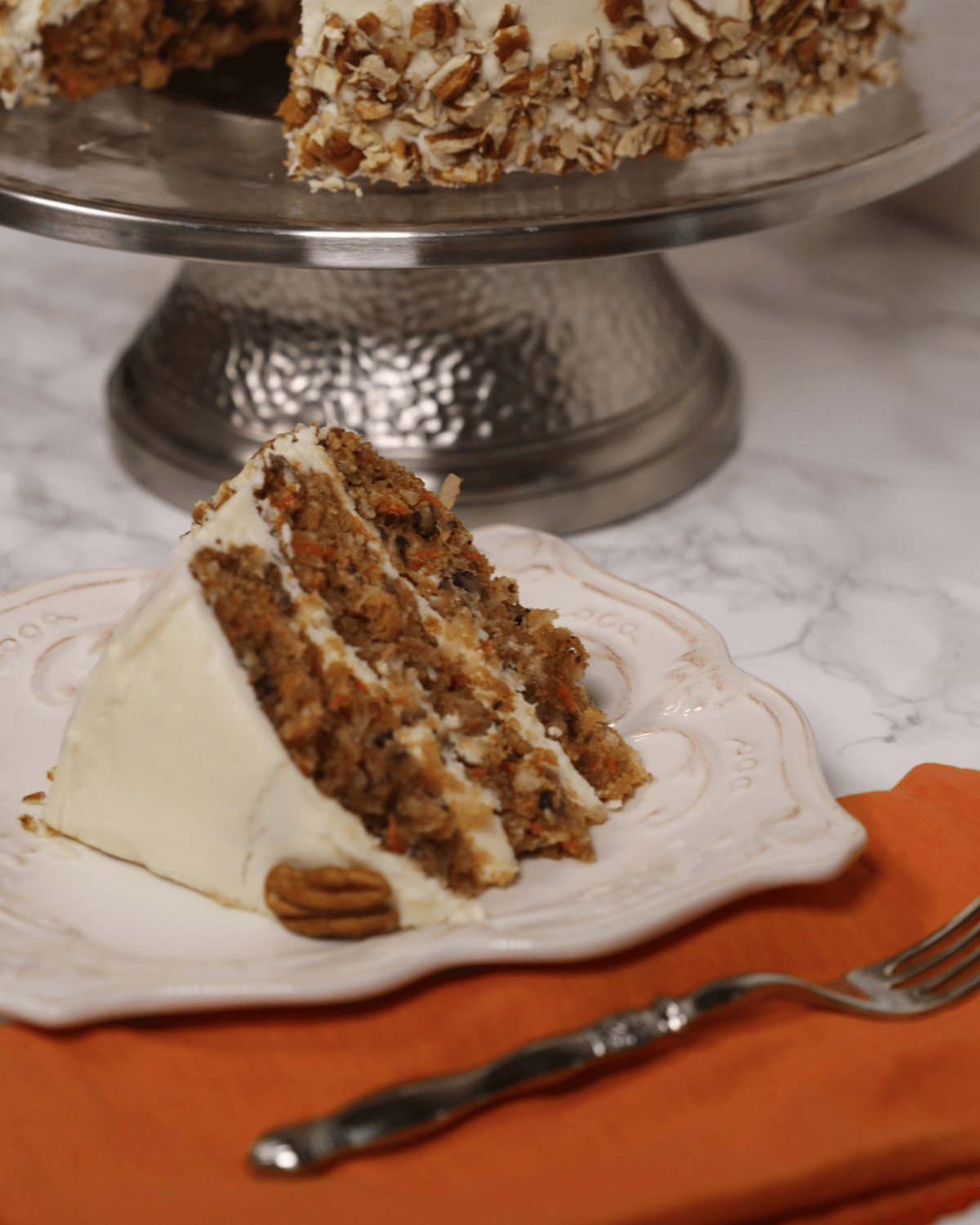 A slice of carrot cake with pineapple, coconut, and cream cheese frosting on a plate, with a cake stand and more cake in the background.