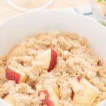 A bowl of apple cinnamon oatmeal mixed with fresh apple slices, placed on a checkered cloth on a table.