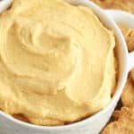 A bowl of creamy pumpkin dip radiates a warm orange hue, nestled amidst an inviting array of crackers on a rustic wooden surface.