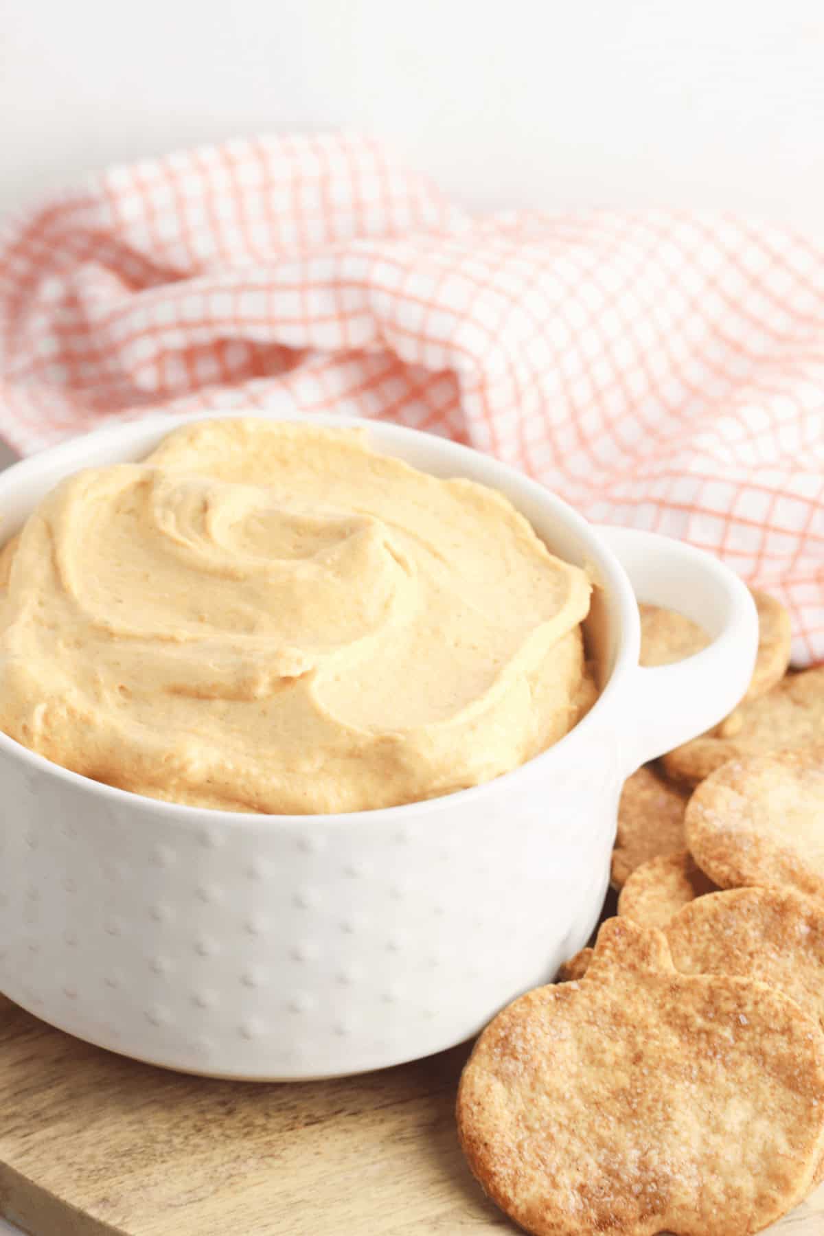 A white bowl of creamy pumpkin dip nestles beside round, golden-brown crackers on a rustic wooden surface, framed by a checkered cloth in the background.