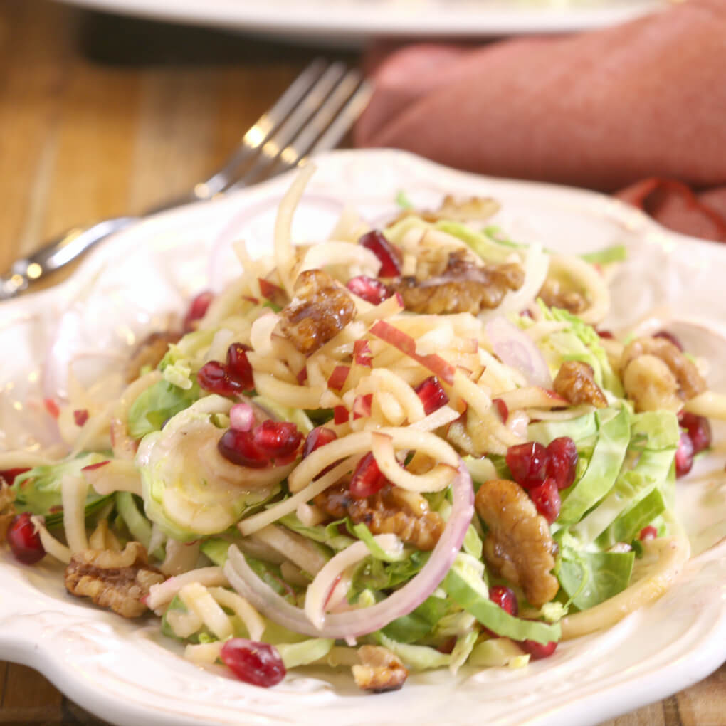 Harvest salad with layers of shaved brussels sprouts, apples, pomegranates, candied walnuts and a pomegranate vinaigrette on a white plate