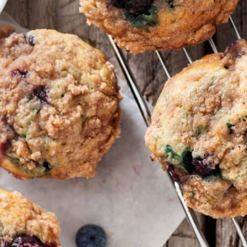 Close-up of freshly baked Blueberry and Buttermilk Muffins with crumb topping on a cooling rack, with a few loose blueberries nearby on a wooden surface.