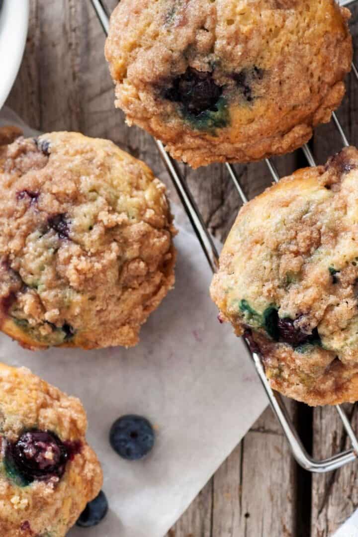 Close-up of freshly baked Blueberry and Buttermilk Muffins with crumb topping on a cooling rack, with a few loose blueberries nearby on a wooden surface.