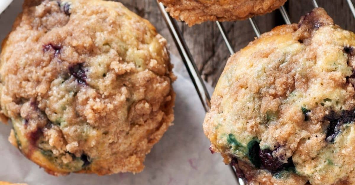 Two Blueberry and Buttermilk Muffins with crumbly streusel topping rest on a cooling rack lined with parchment paper.