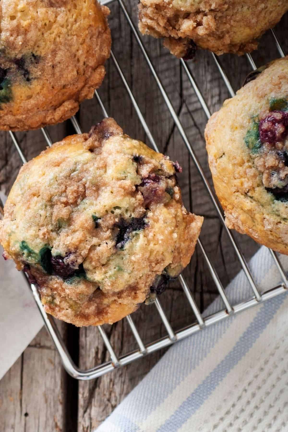 Close-up of Blueberry and Buttermilk Muffins with a crumb topping on a metal cooling rack, set on a rustic wooden surface with a striped cloth nearby.