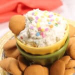 Bowl of funfetti dip and rainbow sprinkles, surrounded by vanilla wafer cookies.