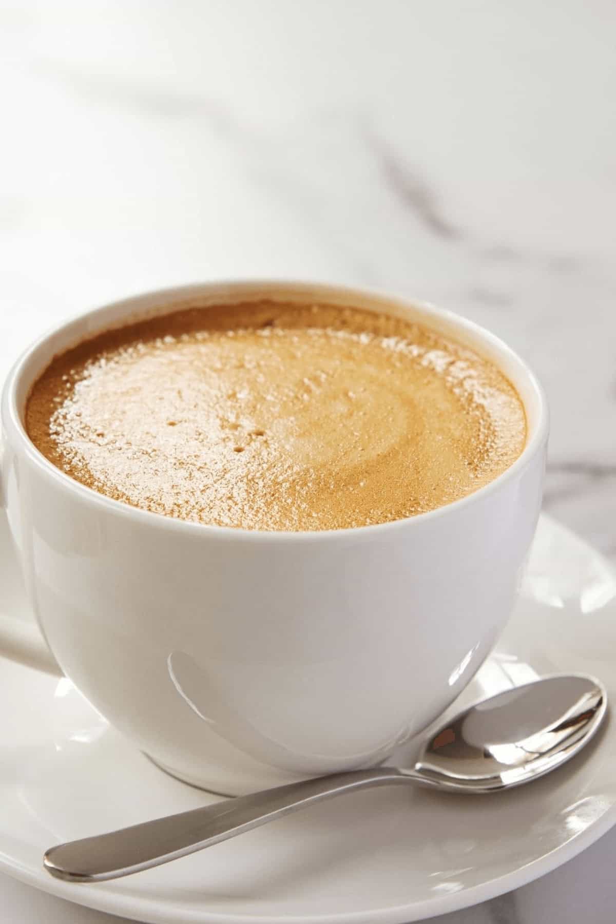 A cup of rich Mocha Latte with frothy crema in a white cup and saucer, accompanied by a silver spoon, rests invitingly on the marble surface.