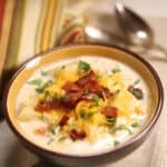A bowl of creamy Chicken Corn Soup topped with shredded cheese, bacon bits, and chopped herbs, with a striped cloth and two spoons in the background.