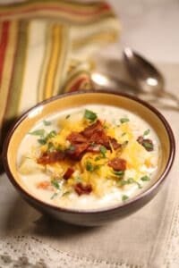 A bowl of creamy Chicken Corn Soup topped with shredded cheese, bacon bits, and chopped herbs, with a striped cloth and two spoons in the background.