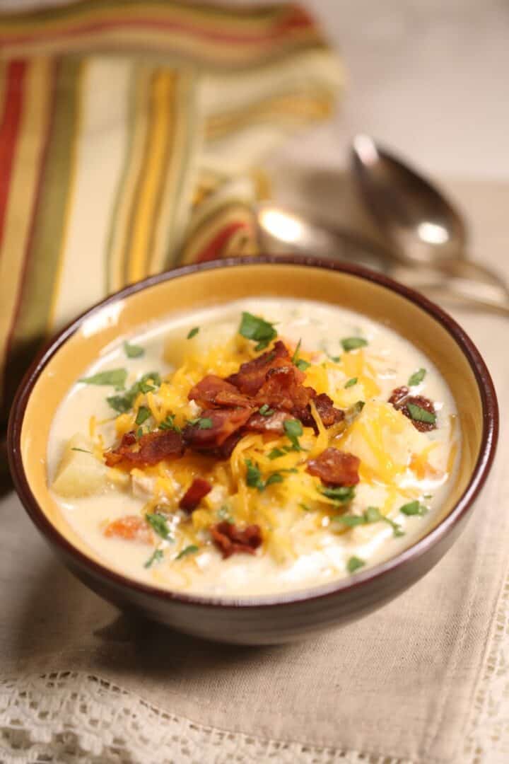 A bowl of creamy Chicken Corn Soup topped with shredded cheese, bacon bits, and chopped herbs, with a striped cloth and two spoons in the background.