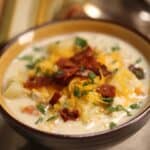 A bowl of creamy Chicken Corn Soup topped with shredded cheese, chopped bacon, and fresh herbs, placed on a cloth napkin with a spoon in the background.