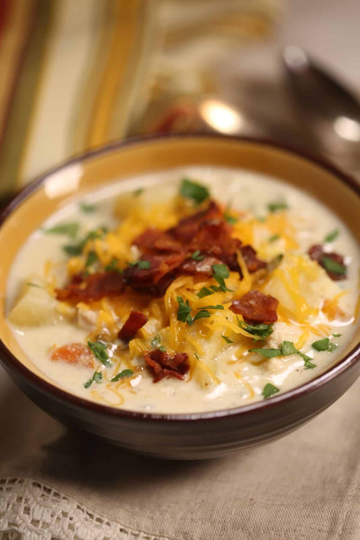 A bowl of creamy Chicken Corn Soup topped with shredded cheese, chopped bacon, and fresh herbs, placed on a cloth napkin with a spoon in the background.