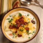 A bowl topped with shredded cheese, bacon bits, and chopped herbs, placed on a cloth napkin beside two spoons and a striped towel.