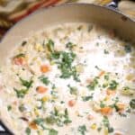 A pot of creamy Chicken Corn Soup with visible chunks of vegetables, corn, and herbs, garnished with chopped parsley, sits on a surface near a striped cloth.