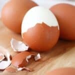 A partially peeled Easy to Peel Hard Boiled Egg sits among whole brown eggs and broken eggshells on a wooden surface.