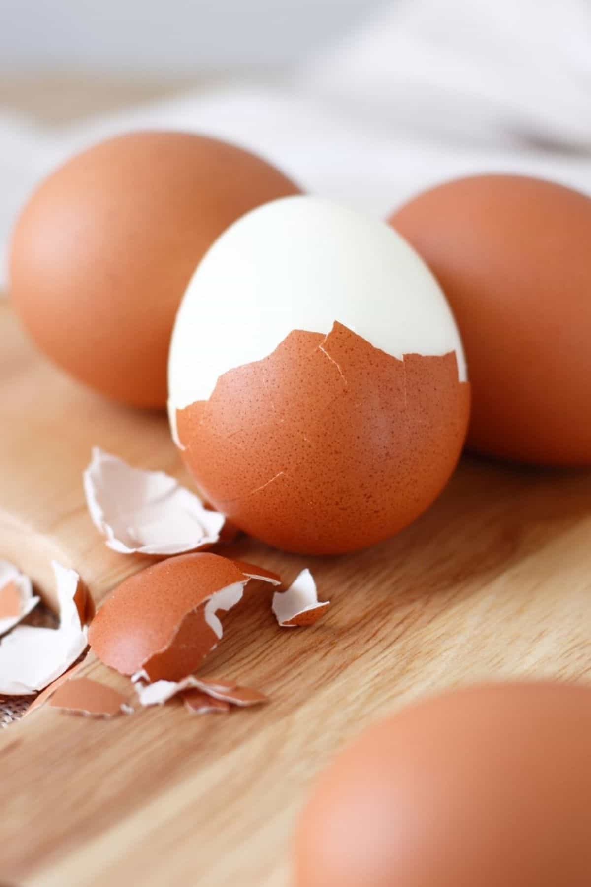 A partially peeled Easy to Peel Hard Boiled Egg sits among whole brown eggs and broken eggshells on a wooden surface.