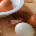 An easy to peel protein and broken eggshells on a wooden surface near a white bowl.