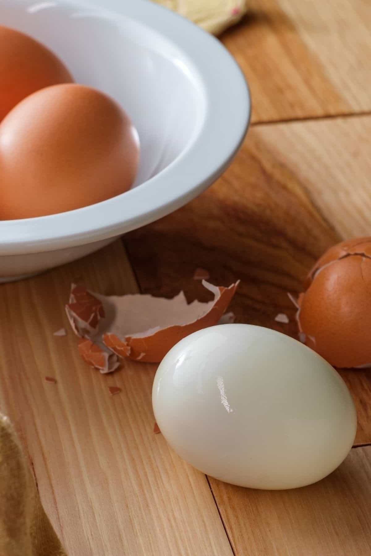 An easy to peel protein and broken eggshells on a wooden surface near a white bowl.