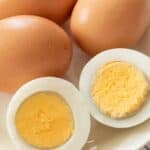 A plate with three whole brown eggs and one Easy to Peel Hard Boiled Egg cut in half, showing the cooked yolk and white.