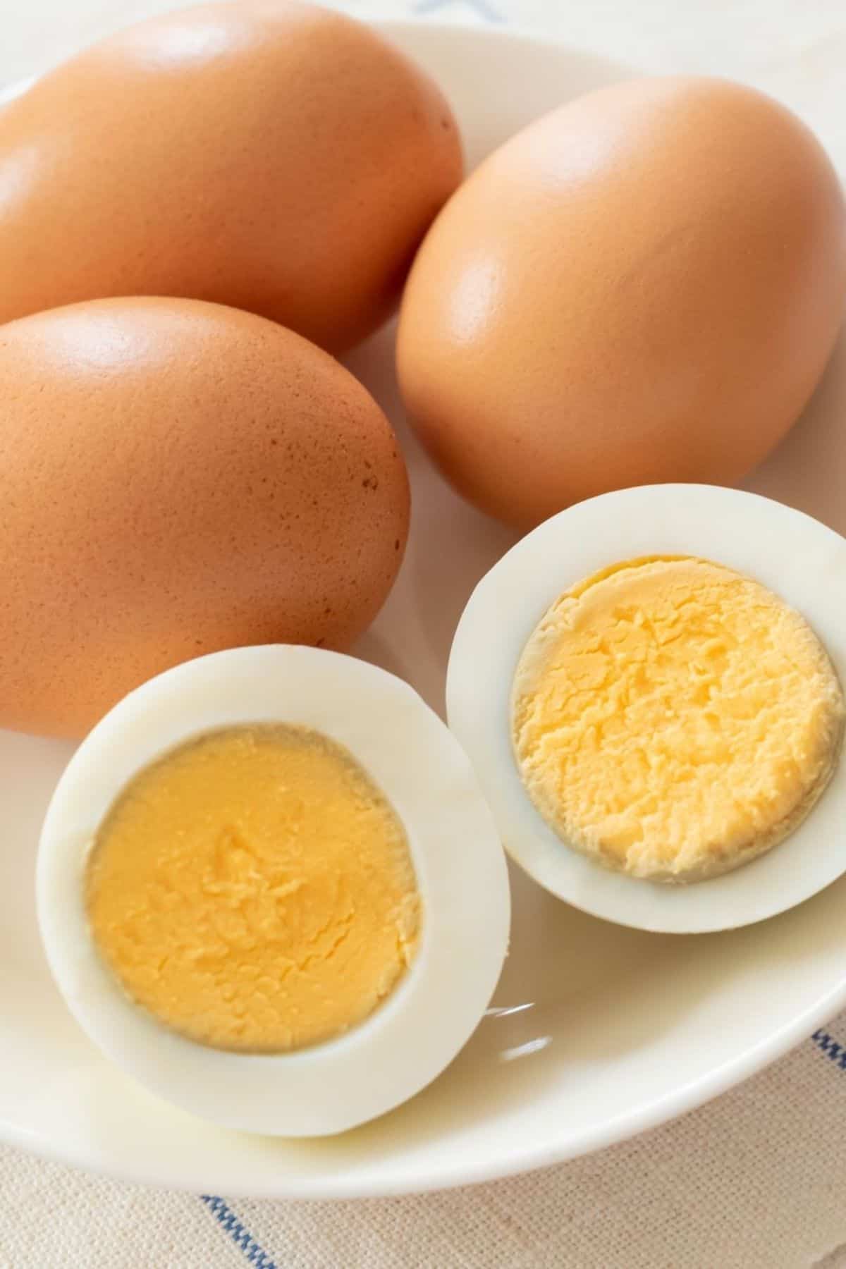 A plate with three whole brown eggs and one Easy to Peel Hard Boiled Egg cut in half, showing the cooked yolk and white.