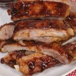 The meat is perfectly tender and flavorful, stacked on a white plate with a red checkered cloth underneath; a bowl of barbecue sauce sits invitingly in the background.