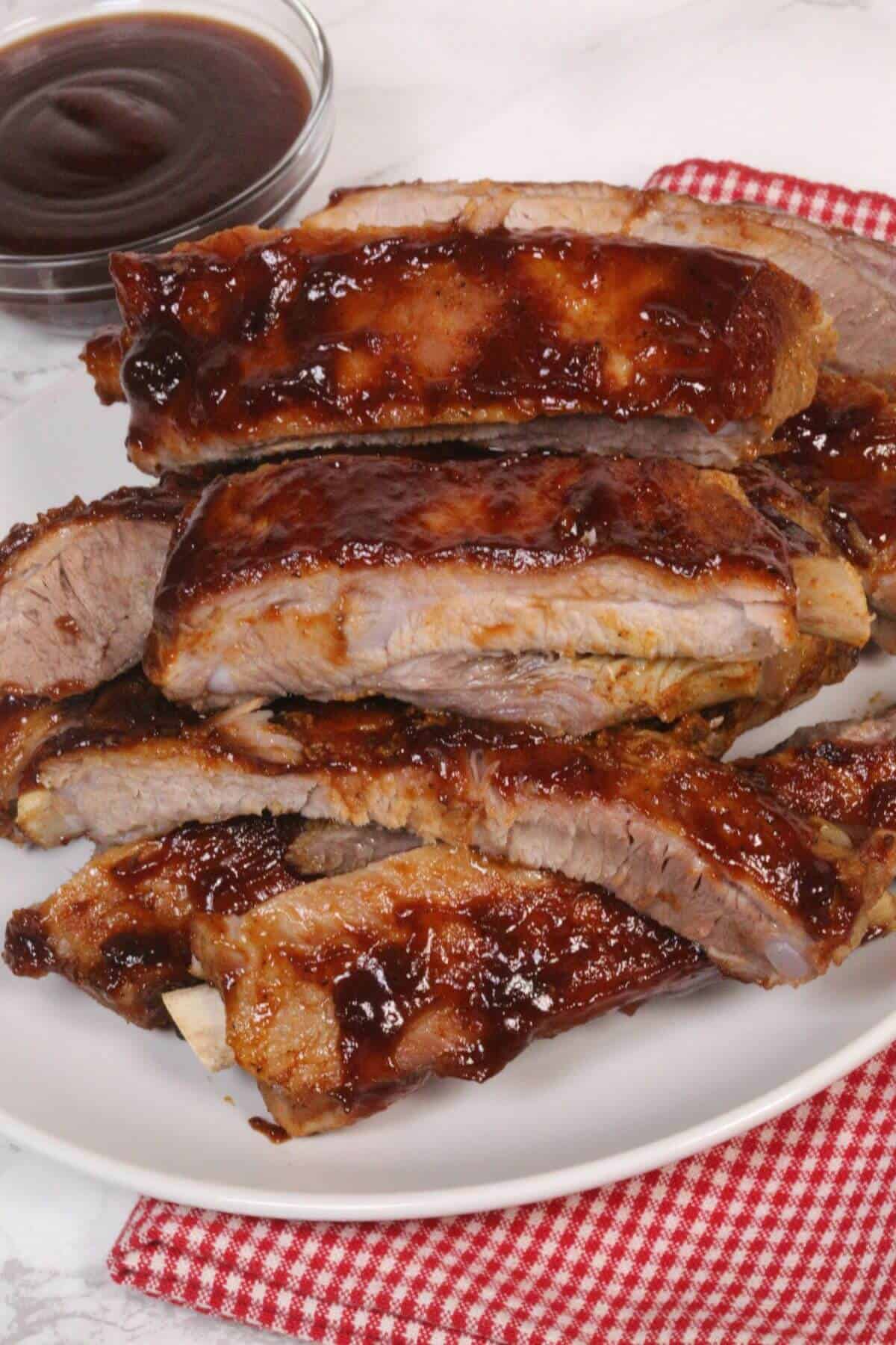The meat is perfectly tender and flavorful, stacked on a white plate with a red checkered cloth underneath; a bowl of barbecue sauce sits invitingly in the background.