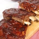 Instant Pot Baby Back Ribs are beautifully barbecued and glazed with sauce, elegantly presented on a wooden board. A bowl of extra sauce waits in the background for those who crave even more flavor.