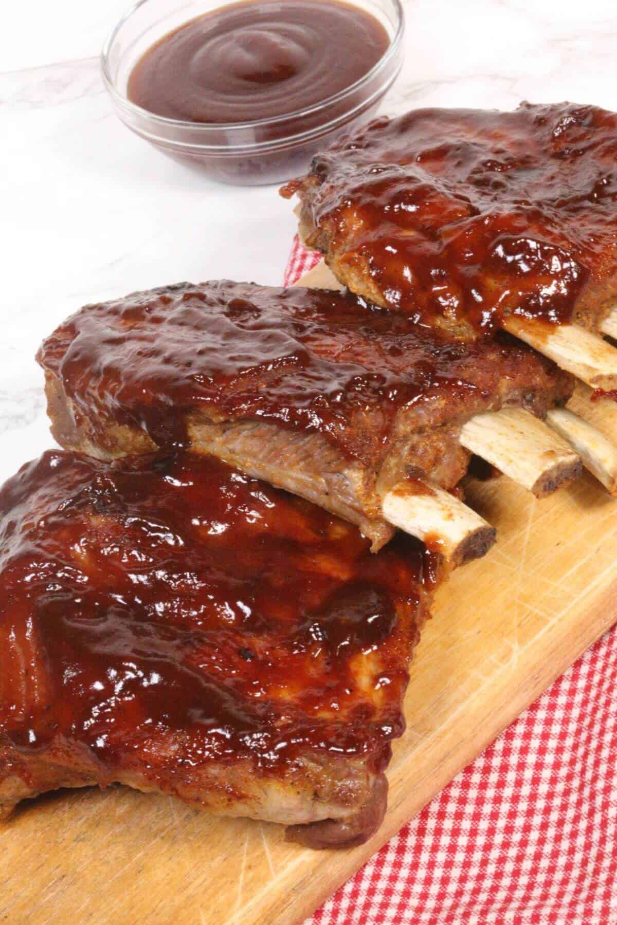Instant Pot Baby Back Ribs are beautifully barbecued and glazed with sauce, elegantly presented on a wooden board. A bowl of extra sauce waits in the background for those who crave even more flavor.