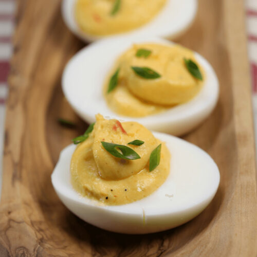 Three spicy deviled eggs on a wooden board, garnished with herbs.