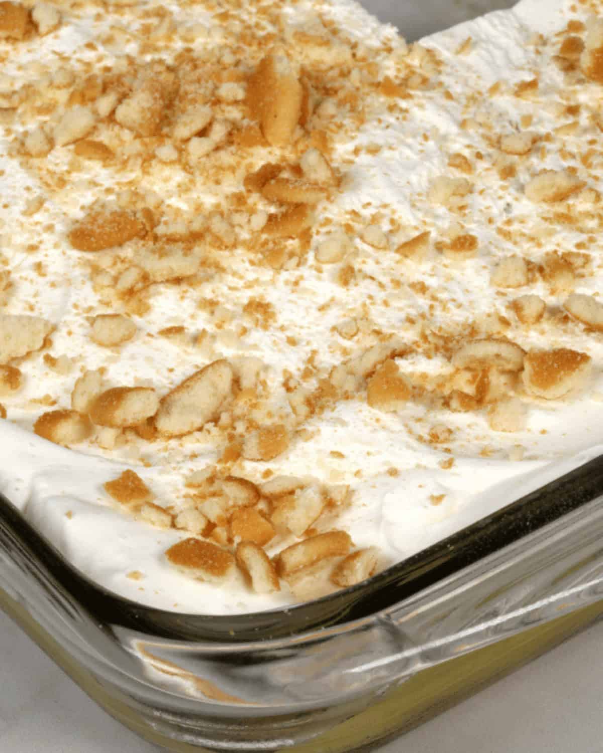 A clear glass dish containing a layered banana cream cake, topped with whipped cream and crushed vanilla wafer cookies.