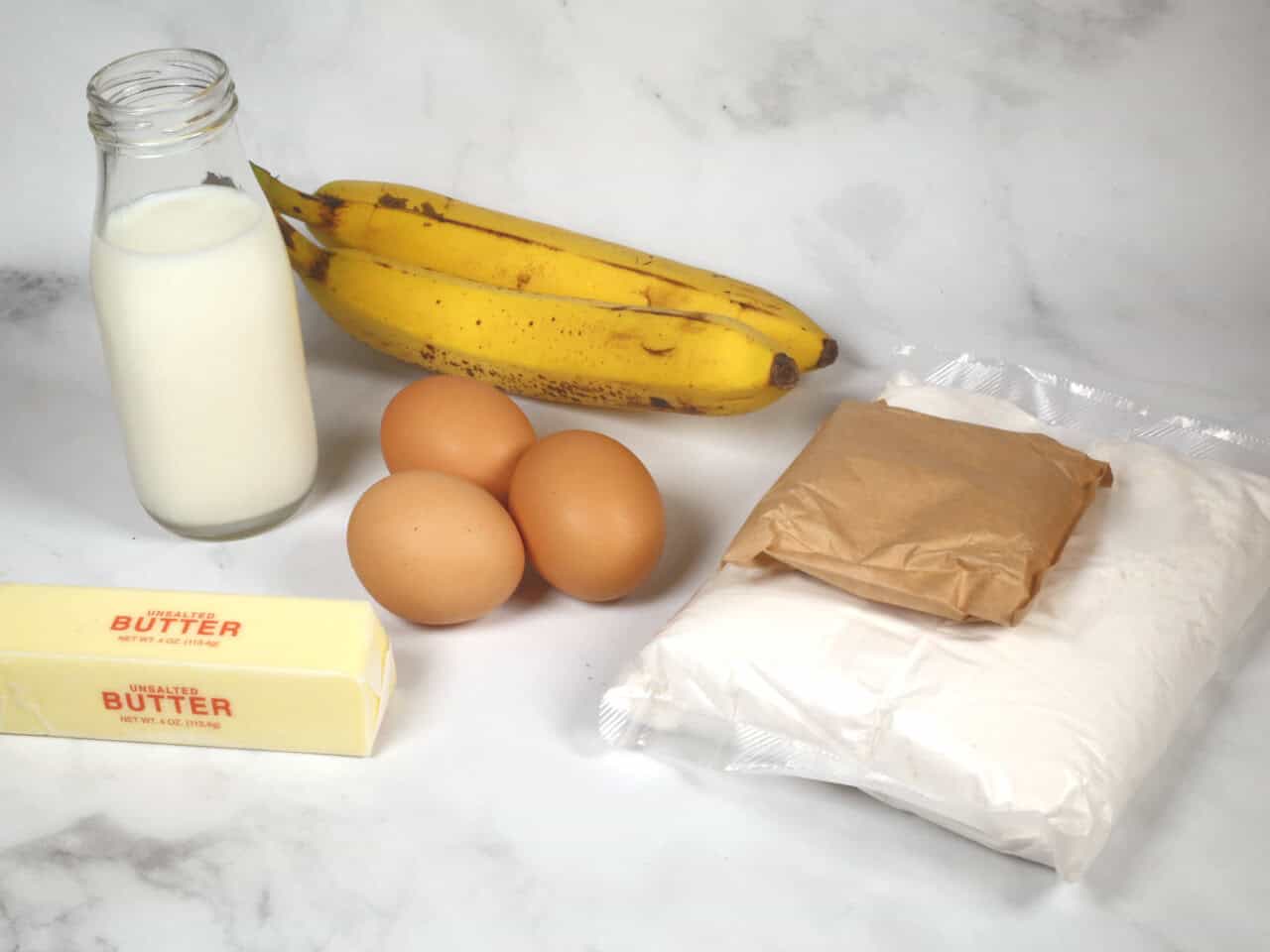 Ingredients for banana cream cake on a marble surface: a bottle of milk, two bananas, three eggs, a stick of butter, and packages of flour and brown sugar.