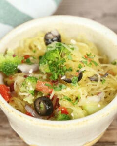 A bowl filled with California Spaghetti Salad, a delightful mix of spaghetti squash, black olives, broccoli, tomatoes, red onions, and garnished with chopped parsley.