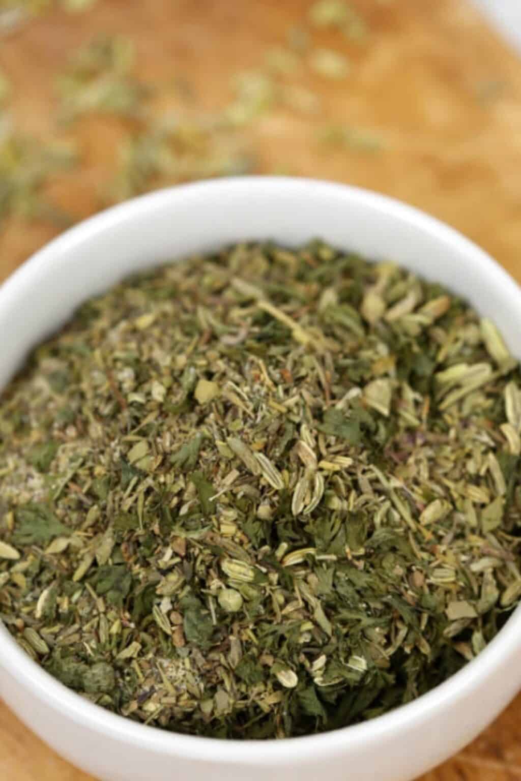 A white bowl filled with Porketta Seasoning, a mixture of dried green and brown herbs, sits on a wooden surface.