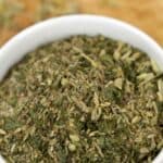 A white bowl filled with Porketta Seasoning, a mixture of dried green and brown herbs, sits on a wooden surface.
