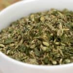 A white bowl filled with a mixture of dried green and brown herbs, known as Porketta Seasoning, sits on a wooden surface.