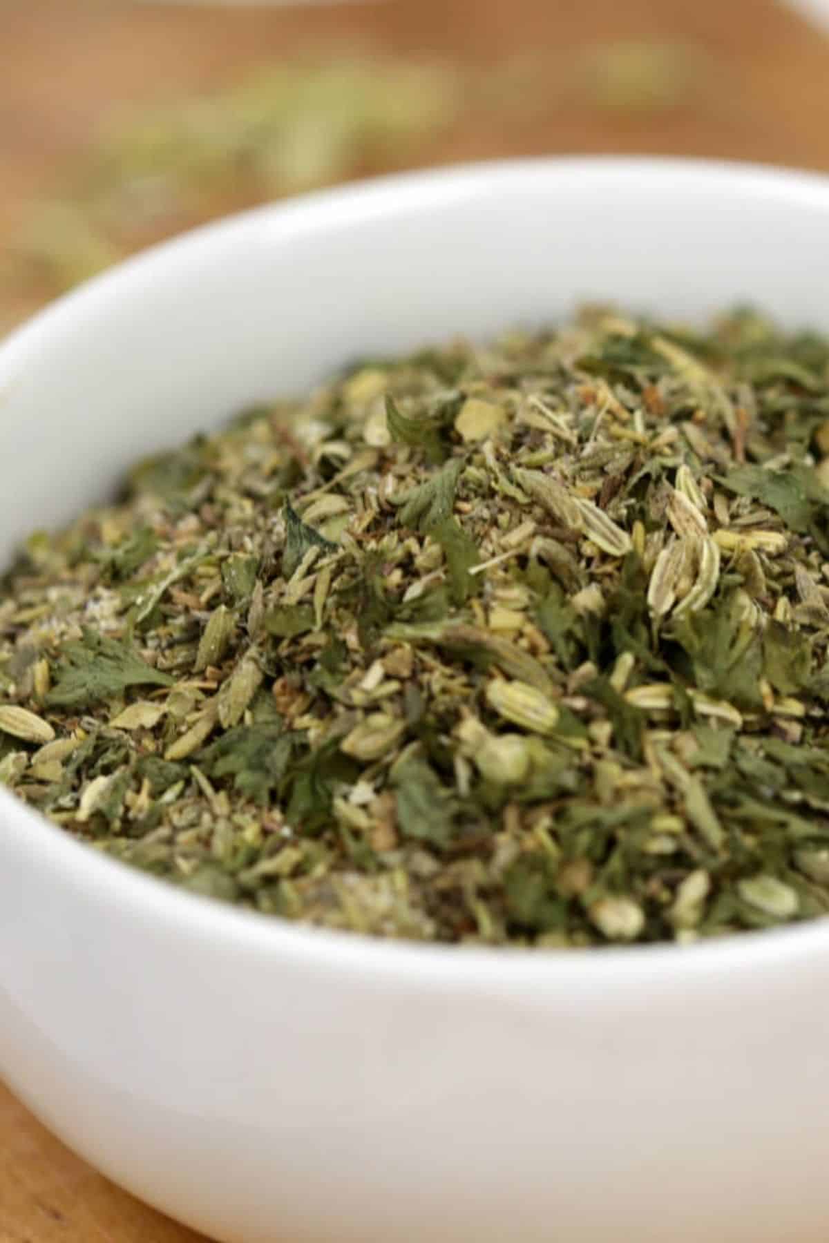 A white bowl filled with a mixture of dried green and brown herbs, known as Porketta Seasoning, sits on a wooden surface.
