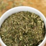 A white bowl filled with a green and brown Porketta Seasoning herb mixture sits on a wooden surface.