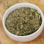 A white bowl filled with a mix of dried Italian herbs and Porketta Seasoning sits on a wooden cutting board.