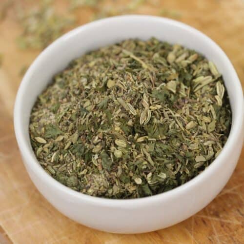A white bowl filled with a mix of dried Italian herbs and Porketta Seasoning sits on a wooden cutting board.