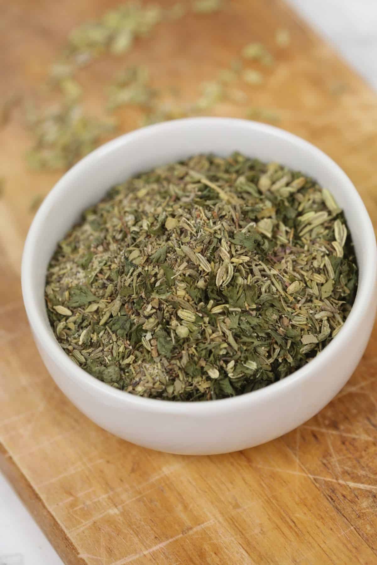 A white bowl filled with a mix of dried Italian herbs and Porketta Seasoning sits on a wooden cutting board.