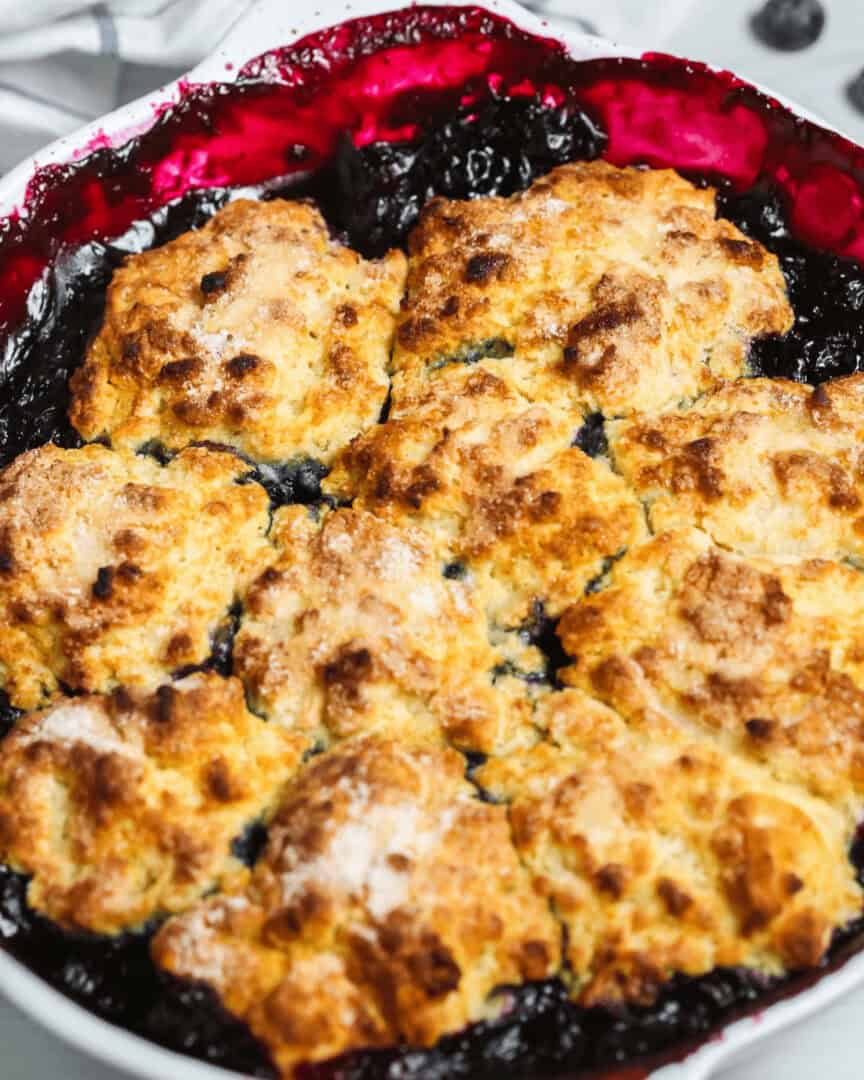 A close-up of a freshly baked, easy blueberry cobbler with golden brown, sugar-topped biscuit crust surrounded by bubbling, dark purple blueberry filling.