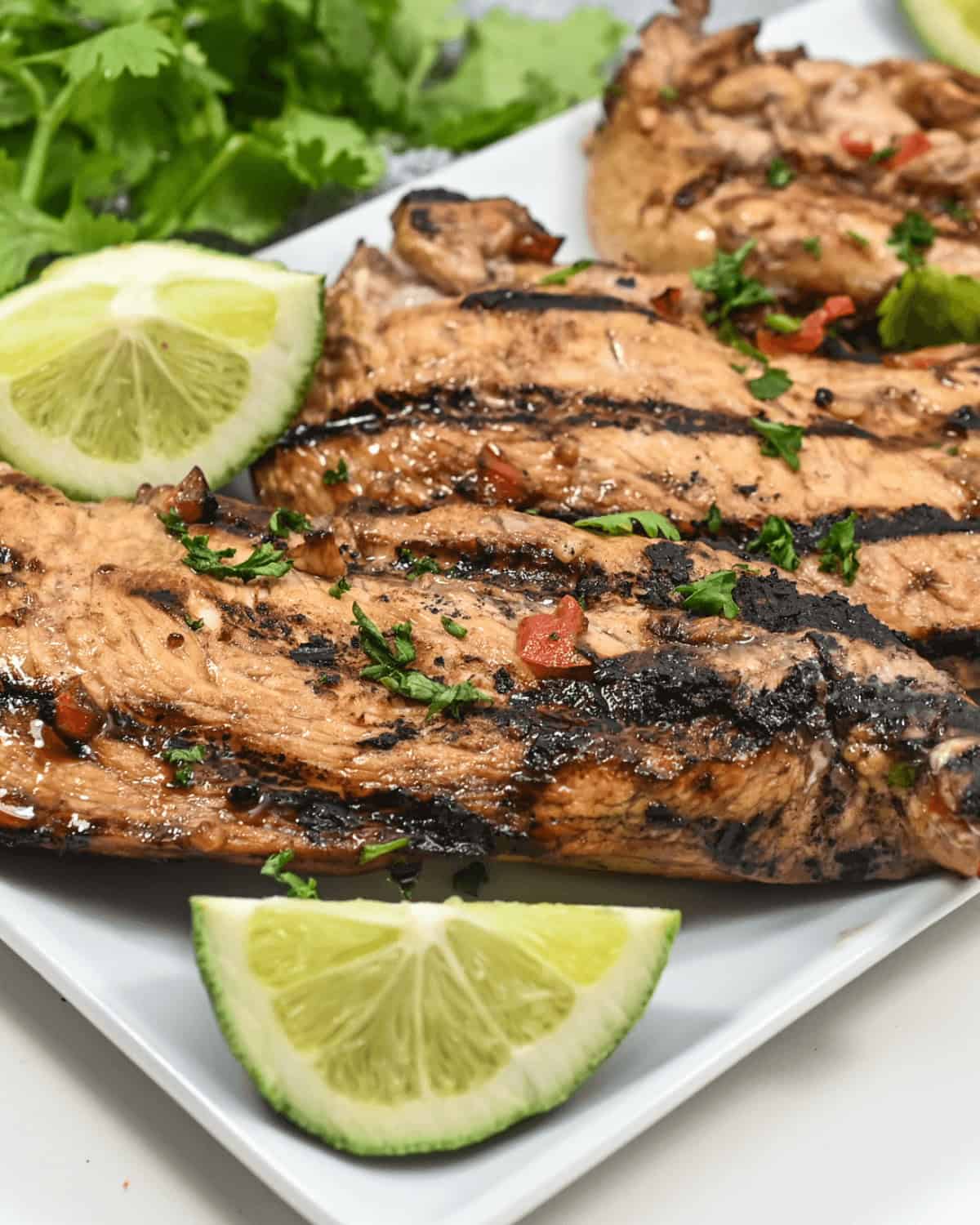 Grilled honey lime chicken breasts garnished with lime wedges and chopped herbs on a white plate.