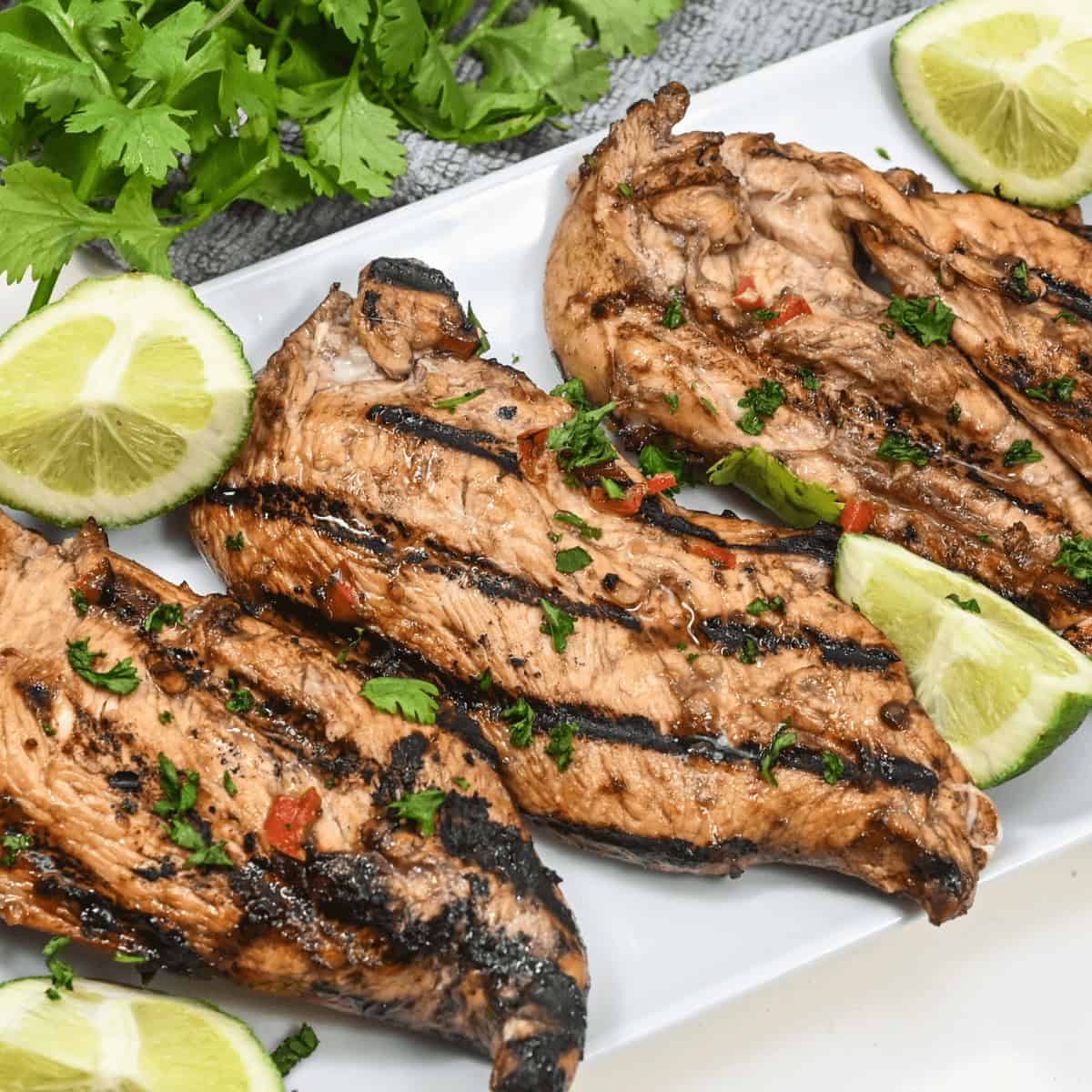 Grilled honey lime chicken breasts garnished with herbs and lime slices on a white plate.