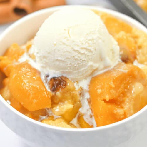 A white bowl filled with peach cobbler topped with a scoop of vanilla ice cream. Cinnamon sticks are visible in the background, reminiscent of an Easy Blueberry Cobbler.