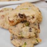 A stack of three with visible chocolate chips and green pieces, one with a bite taken out, sits on a white plate with more in the background.